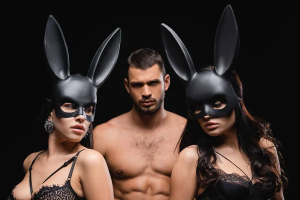 Donne appassionate in biancheria intima e maschere coniglietto vicino uomo sexy su sfondo sfocato isolato su nero — Foto stock