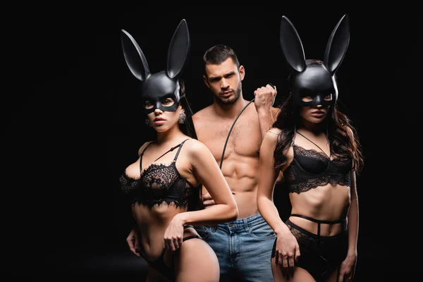 Homme dominant tenant fouet fouet près de femmes chaudes dans des masques de lapin sur fond noir — Photo de stock