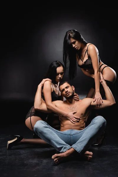 Сексуальный мужчина сидит со скрещенными ногами и смотрит в камеру рядом с чувственными женщинами на черном фоне — стоковое фото