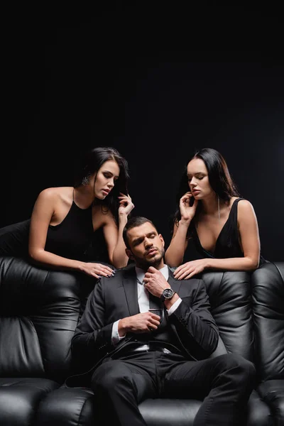 Businessman adjusting tie while sitting on leather couch near sexy women standing isolated on black — Stock Photo