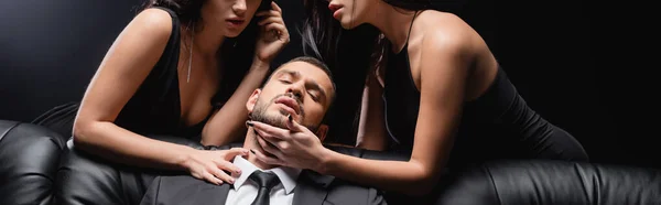 Leidenschaftliche Frauen berühren Geschäftsmann auf Ledercouch auf schwarz, Banner — Stockfoto