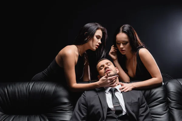 Passionate, brunette women seducing businessman sitting on leather couch on black — Stock Photo