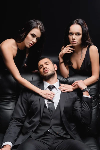 Élégant homme en costume assis sur canapé en cuir près de femmes sexy isolé sur noir — Photo de stock