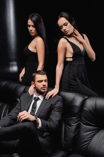 Morena mujeres en elegantes vestidos tocando hombros de hombre de negocios sentado en el sofá de cuero en negro — Stock Photo