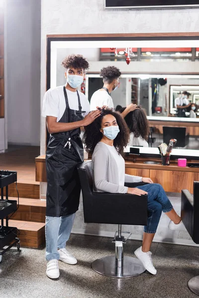 Peluquero afroamericano en máscara protectora mirando a la cámara cerca de la mujer joven - foto de stock