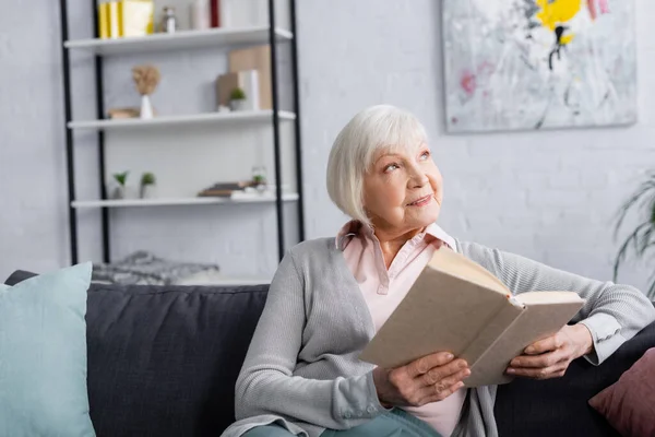 Lächelnde ältere Frau hält Buch in der Hand und schaut zu Hause auf — Stockfoto