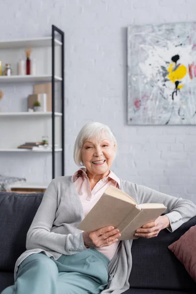 Mujer mayor sonriendo a la cámara mientras sostiene el libro en el sofá - foto de stock