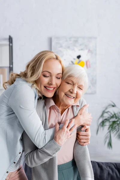 Glückliche Frau umarmt grauhaarige Mutter mit geschlossenen Augen im Wohnzimmer — Stockfoto