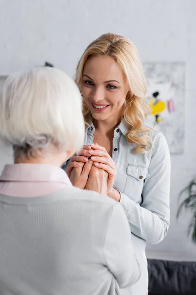 Lächelnde Frau hält die Hände der grauhaarigen Mutter im Wohnzimmer — Stockfoto