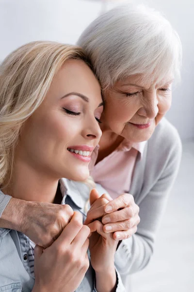 Fröhliche Frau hält ältere Mutter mit geschlossenen Augen zu Hause an der Hand — Stockfoto