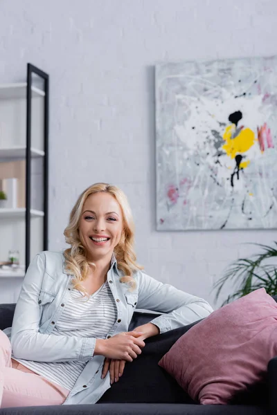 Femme blonde souriant à la caméra près de l'oreiller sur le canapé à la maison — Photo de stock