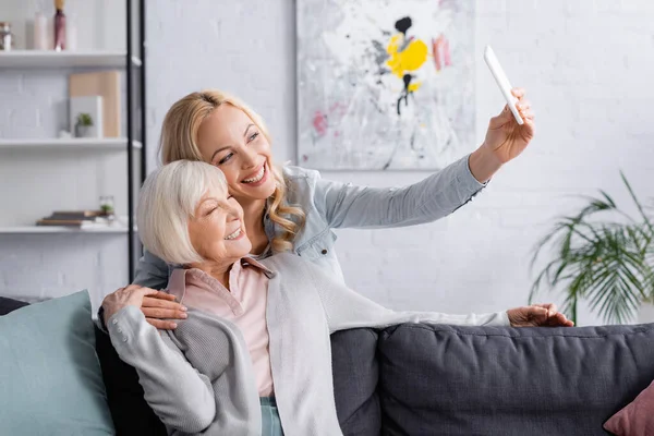 Donna sorridente che si fa selfie con un genitore anziano a casa — Foto stock