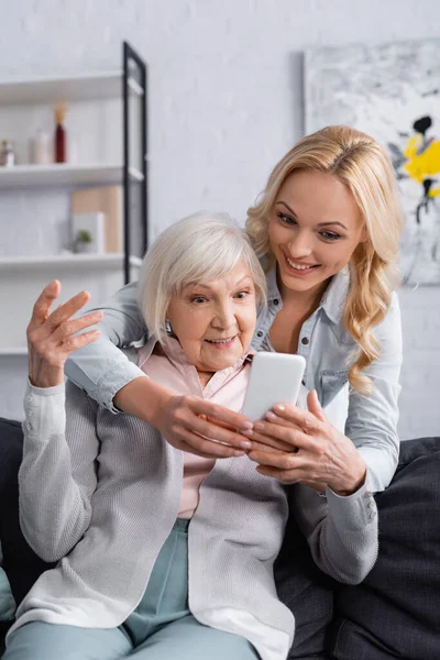 Улыбающаяся женщина, использующая мобильный телефон рядом с пожилой матерью в гостиной — стоковое фото