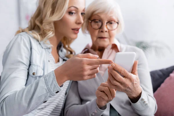 Frau auf verschwommenem Hintergrund zeigt mit Finger auf Smartphone in den Händen der Mutter — Stockfoto