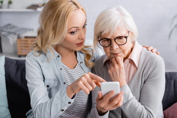 Woman using smartphone near pensive mother in eyeglasses — Stock Photo