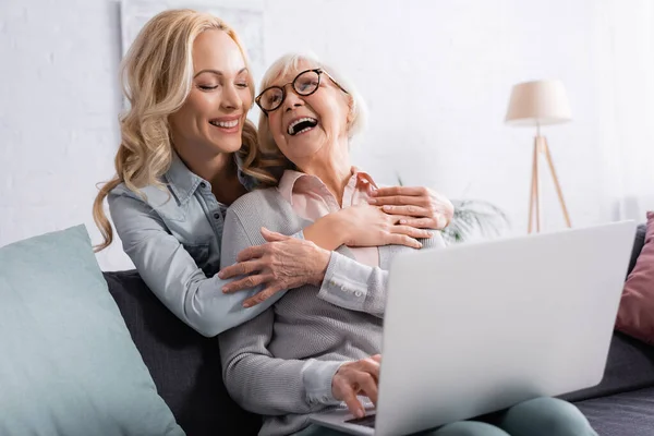 Donna allegra che abbraccia la madre anziana con laptop sfocato a casa — Foto stock