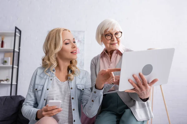 Smiling woman with smartphone pointing at laptop near senior mother at home — Stock Photo