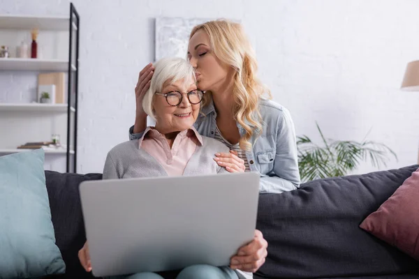 Erwachsene Frau küsst lächelnde Mutter mit Laptop auf verschwommenem Vordergrund — Stockfoto