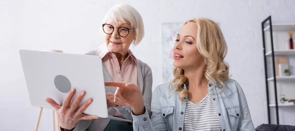 Mulher apontando para laptop perto de mãe idosa em casa, banner — Fotografia de Stock