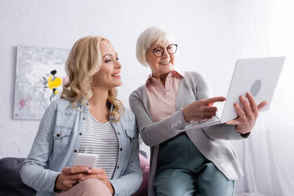 Mujer anciana alegre señalando con el dedo a la computadora portátil cerca de hija sonriente con teléfono inteligente - foto de stock