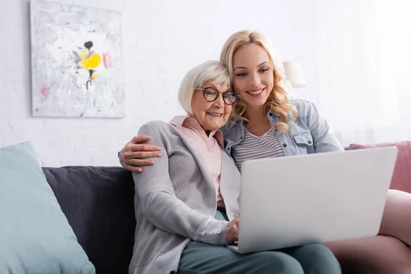 Mujer sonriente abrazando a la madre mayor mientras usa el ordenador portátil en la sala de estar - foto de stock