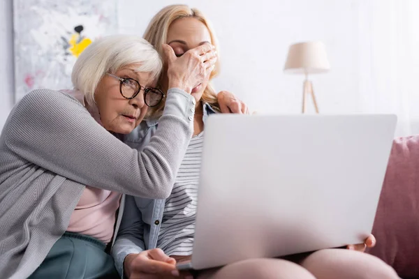 Seniorin verdeckt Augen ihrer Tochter in der Nähe von Laptop auf verschwommenem Vordergrund zu Hause — Stockfoto