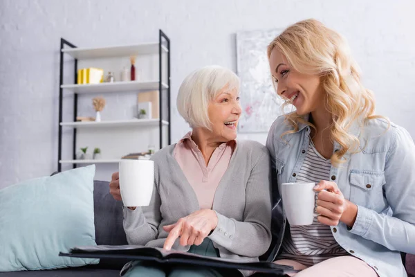Старшая женщина указывает на фотоальбом рядом с улыбающейся дочерью с чашкой — стоковое фото