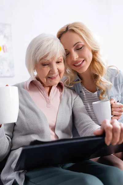 Lächelnde Seniorin mit Tasse hält Fotoalbum neben Tochter — Stockfoto