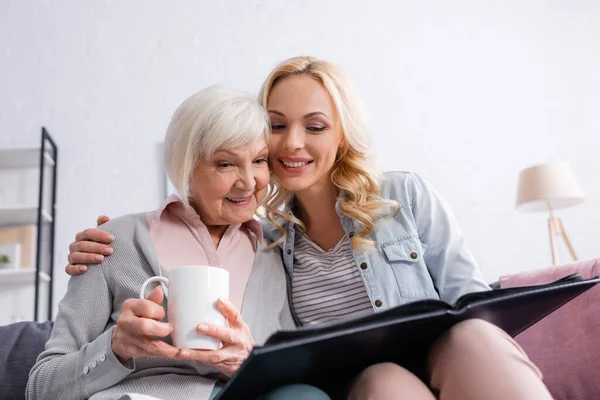 Mujer positiva abrazando a la madre con taza mientras mira el álbum de fotos - foto de stock