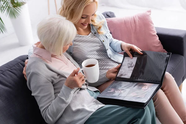 Smiling women with tea looking at photos in album in living room — Stock Photo