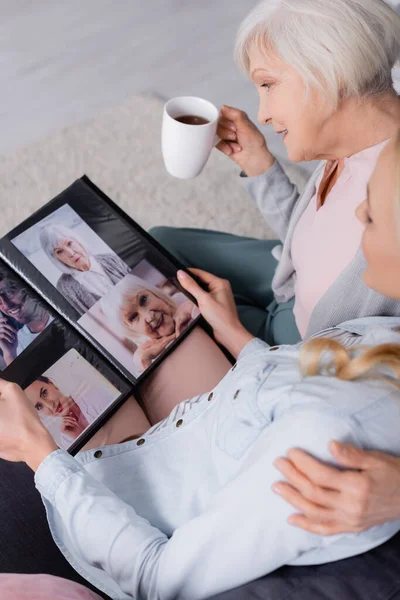 Femme âgée souriante avec tasse regardant des photos dans l'album près de fille floue — Photo de stock