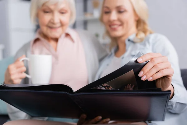 Fotoalbum in Händen einer verschwommenen Frau nahe der Mutter mit Tasse — Stockfoto