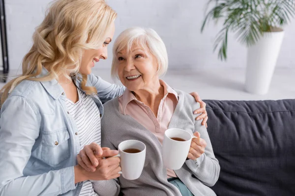Donna sorridente con tè che abbraccia la madre anziana a casa — Foto stock