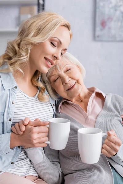 Sonriente anciana sosteniendo la taza mientras está sentada cerca de una hija adulta - foto de stock