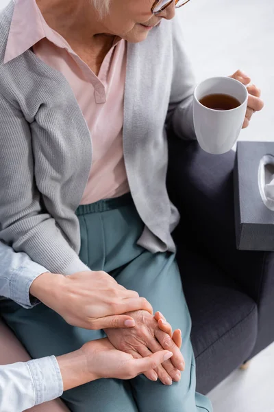 Vista recortada de la mujer mayor sosteniendo la taza de té cerca de la hija sosteniendo la mano - foto de stock
