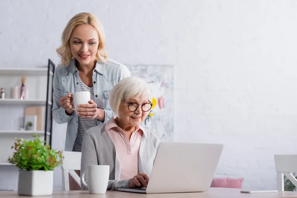 Smiling woman with cup sanding near mother using laptop — Stock Photo