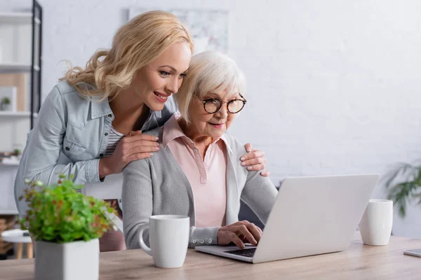 Smiling woman hugging mother using laptop near cups — Stock Photo