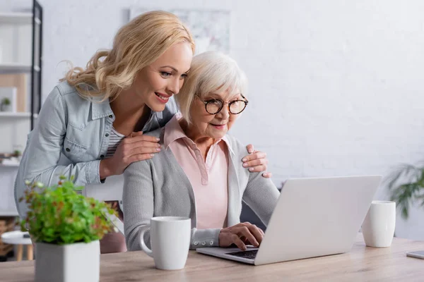 Donna sorridente che abbraccia la madre usando il computer portatile vicino alle tazze — Foto stock