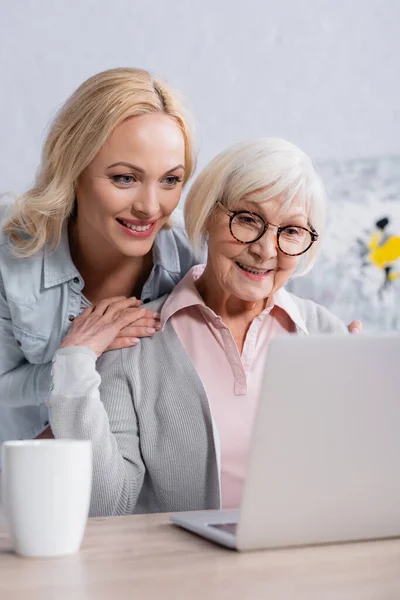 Mujer alegre abrazando a la madre mayor cerca de la computadora portátil y la taza en primer plano borrosa - foto de stock