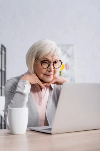 Старшая женщина в очках смотрит на ноутбук рядом с чашкой — стоковое фото