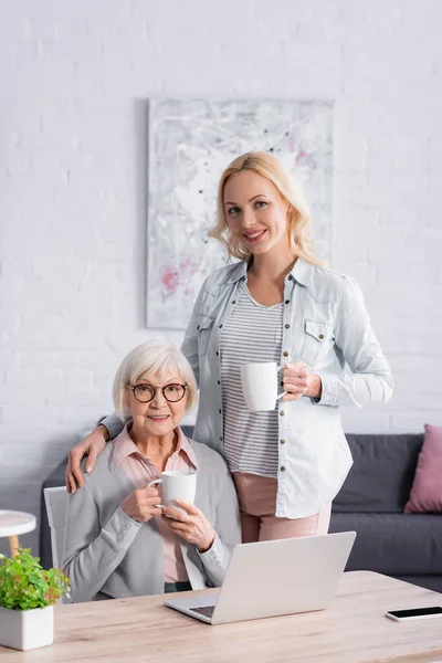 Fröhliche Frau mit Tasse neben Mutter und Spielzeug auf Tisch — Stockfoto