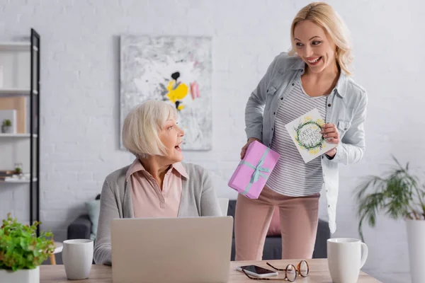 Mujer sonriente sosteniendo tarjeta de felicitación y regalo cerca de la madre y los dispositivos - foto de stock