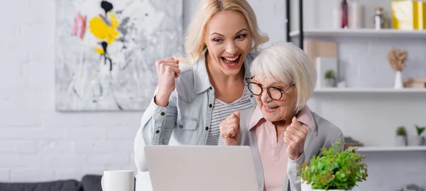 Mujer positiva mostrando sí gesto cerca de la computadora portátil y la madre anciana, bandera - foto de stock