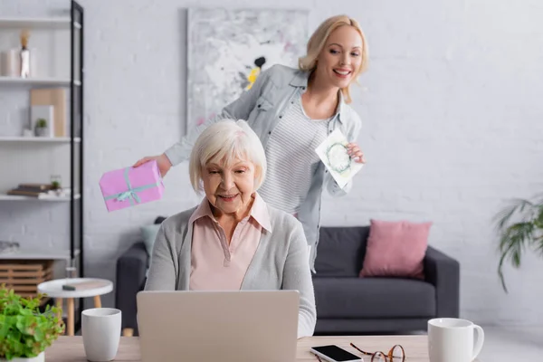 Donna anziana utilizzando laptop vicino figlia con presente e biglietto di auguri su sfondo sfocato — Foto stock