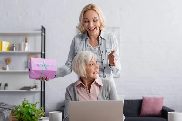 Mujer alegre con caja de regalo de pie cerca de la madre mayor y el ordenador portátil - foto de stock
