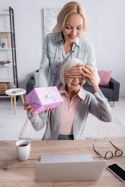 Femme souriante couvrant les yeux de la mère tout en tenant présent près de l'ordinateur portable et le thé sur la table — Photo de stock