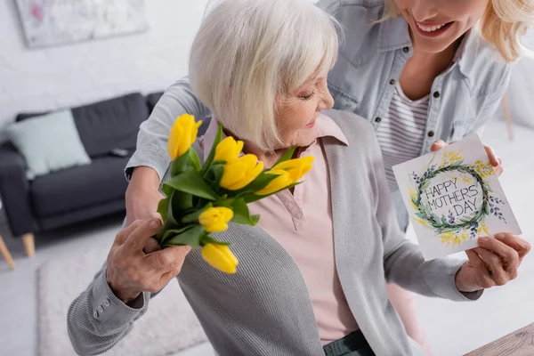 Donna sorridente che tiene biglietto di auguri e fiori vicino genitore anziano — Foto stock