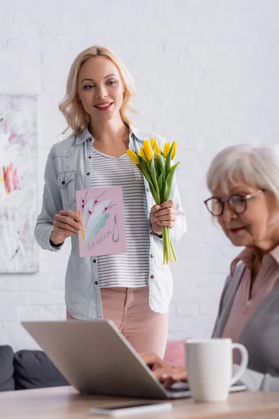 Donna sorridente che tiene biglietto di auguri con 8 lettere di marcia e fiori vicino alla madre anziana utilizzando il computer portatile — Foto stock