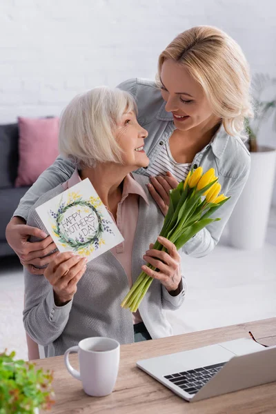 Senior woman holding flowers and greeting card with happy mothers day lettering near daughter and laptop — Stock Photo