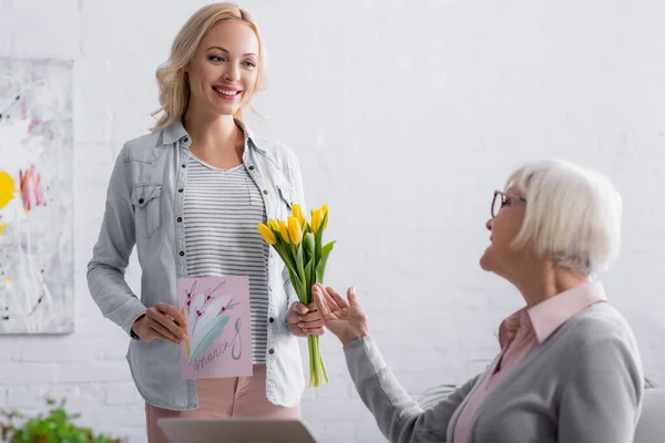 Fröhliche Frau mit Grußkarte und Tulpen neben älterer Mutter im verschwommenen Vordergrund — Stockfoto